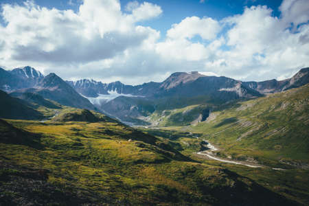 Scenic green blue alpine landscape with mountain river in highland valley in sunlight and big glacier under cloudy sky. Shadow of clouds on green mountain valley. Clouds shadow on rocks and hills.の写真素材