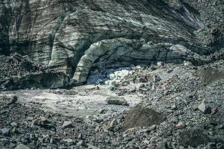Scenic mountain landscape with hikers among ice blocks of cracked glacier. Beautiful scenery with tourists with backpacks among large moraines and ice chunks of big glacier with scratches in mountainsの写真素材