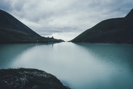 Dark atmospheric landscape with orange tent near mountain lake in highland valley under cloudy sky in dark time. Ripples on mountain lake water in dusk. Dark scenery with water ripple on highland lakeの写真素材