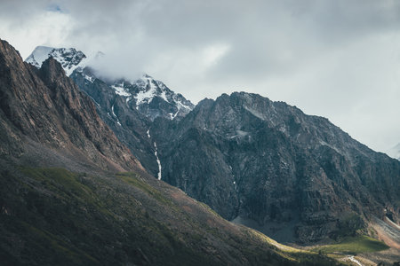Awesome mountain landscape with snowy mountains in low clouds. Rocky top and snowy top. Atmospheric view to great rocky mountains in cloudy sky. Gloomy scenery with high mountains in overcast weather.の写真素材