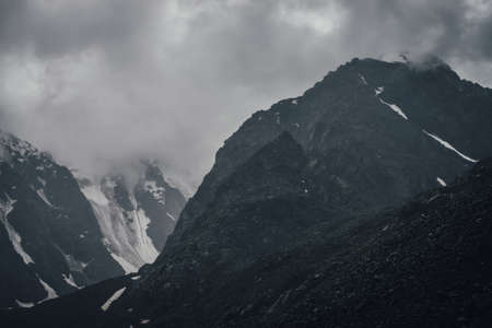 Dark atmospheric mountain landscape with glacier on black rocks in lead gray cloudy sky. Snowy mountains in low clouds in rainy weather. Gloomy landscape with black rocky mountains with snow in fog.の写真素材