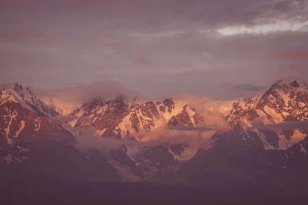 Scenic mountain landscape with great snowy mountain range lit by dawn sun among low clouds. Awesome alpine scenery with high mountain ridge at sunset or at sunrise. Big glacier on top in vintage colorの写真素材