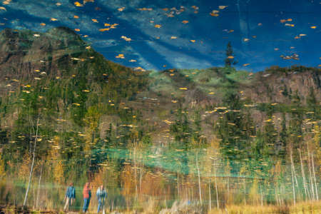 Yellow autumn leaves on clear water surface of mountain lake with reflection of people and golden trees in sunshine. Bright nature background of beautiful turquoise lake in autumn colors in fall time.の写真素材