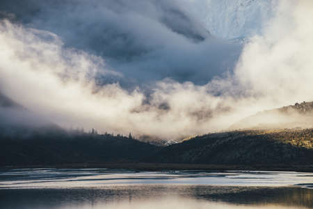 Mountain landscape with shiny lake and silhouettes of coniferous trees on dark rocks with view to high snowy mountain wall in low clouds in golden sunshine. Layered scenery with shadow and sunlight.の写真素材