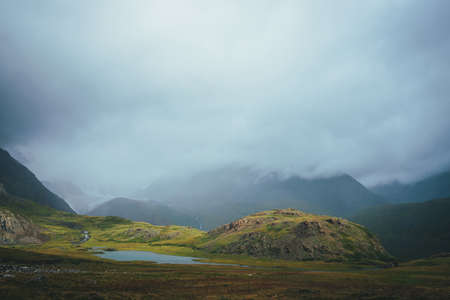 Dramatic alpine landscape with beautiful mountain lake in green valley among great rocks and high mountains in overcast weather. Awesome view to lake and glacier in low clouds. Atmospheric scenery.の写真素材