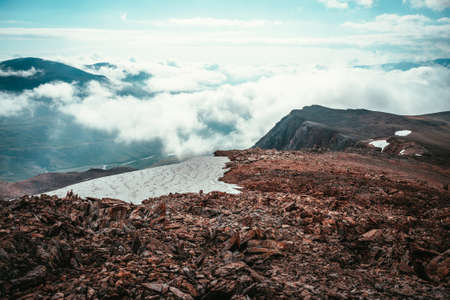 Epic view from precipice edge with snow cornice above clouds to mountain valley in low clouds. Surrealist scenery with low clouds in highland valley. Surreal alpine landscape with high rocky mountainsの写真素材