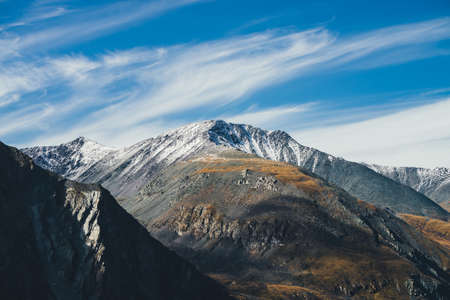 Colorful alpine landscape with great mountain in autumn colors with snow on peak in sunshine under cirrus clouds in blue sky. Picturesque autumn scenery with sunlit rocks and snow-covered mountain topの写真素材