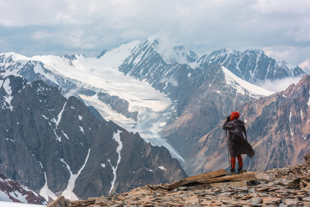 Hiker at very high altitude with view to long glacier tongue and high snow mountains under cloudy sky. Man on windy stone hill against huge mountains with low clouds. Man and majestic mountains natureの写真素材