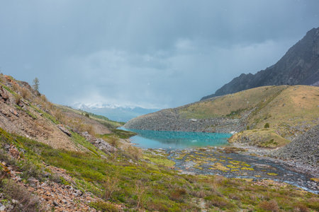 Colorful landscape with azure mountain lake among green hills and sharp rocks against snowy mountain range under cloudy sky during snowfall. Vivid alpine lake in sunlit picturesque mountain valley.の写真素材