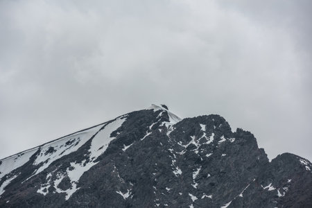 Monochrome scenery with high mountain peak with snow cornice in gray cloudy sky at overcast. Dark...