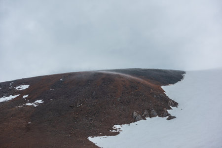 Dark atmospheric landscape with low cloud on high rocky mountain near snow-capped top at overcast. Gloomy alpine scenery with fog on rocks and snowbound mountain under gray rainy sky at cloudy weatherの写真素材