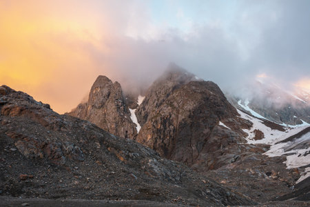 Scenic mountain landscape with sharp rocks in low clouds in orange sunrise colors. Awesome view to...