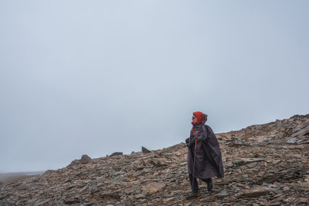 Tourist on stone hill at very high altitude under gray cloudy sky. Hiker with trekking poles and camera on rocky mountain in overcast weather. Man in high mountains among sharp rocks and sharp stones.の写真素材