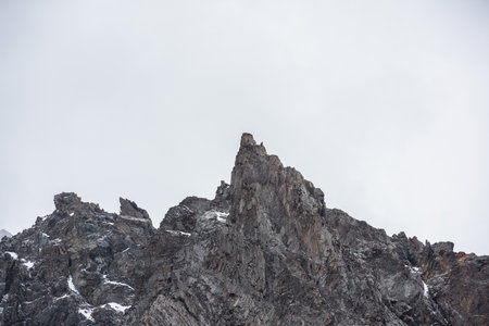Gloomy mountain landscape with high sharp rockies with snow in gray rainy sky. Closeup of sharp rocks and peaked top in gray cloudy sky. Bleak overcast scenery with mountain range with pointy peak.の写真素材