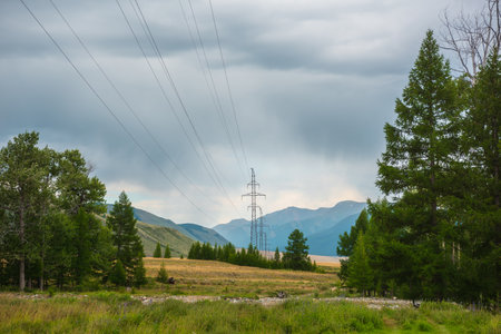 Dramatic mountain landscape with tall tower of power lines under cloudy sky. Atmospheric alpine scenery with electric tower with wires against high mountains in overcast. Electricity in mountains.の写真素材