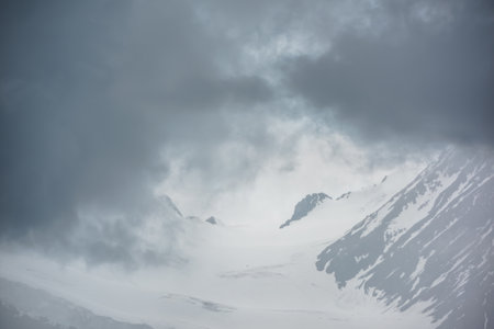Atmospheric mountain landscape with fuzzy silhouettes of rocks on glacier in low clouds during rain. Dramatic view to large glacier tongue in snow mountains blurred in rain haze in gray low clouds.の写真素材