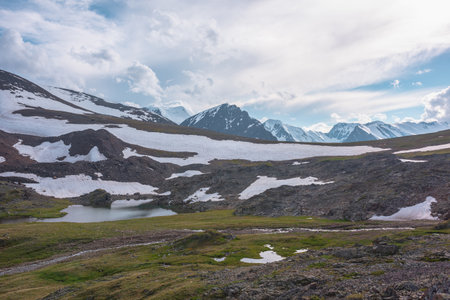 Colorful landscape with mountain lake among hills and rocks against sunlit high snowy mountain range under cloudy sky in changeable weather. Awesome sunny scenery with lake and snowy mountain top.の写真素材
