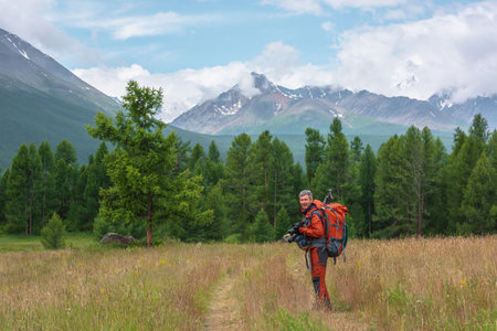 Traveling man in red with big backpack on way to snowy mountain range in low clouds. Backpacker walks through forest to large mountains under cloudy sky. Dramatic landscape with tourist in mountains.の写真素材