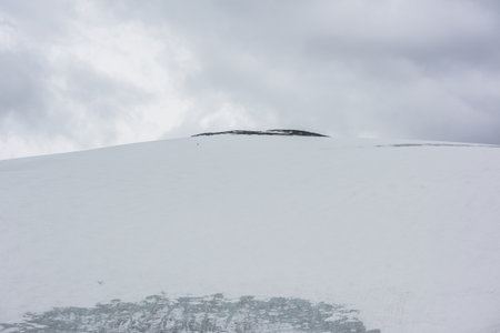 Dramatic landscape with huge snowy mountain dome with rocky top under gray cloudy sky. Awesome scenery with high snow mountain in dome shape in center at overcast weather. Dome shaped mountain top.の写真素材