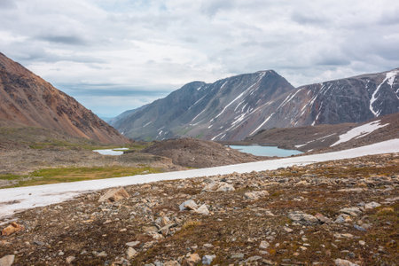 Dramatic landscape with glacier and stone hill between two mountain lakes in sunlight under cloudy sky. Two small lake in high mountain valley with view to mountain vastness at changeable weather.の写真素材