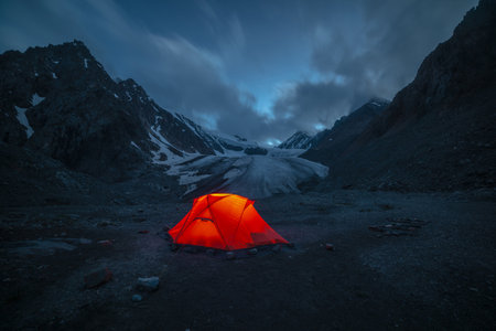 Awesome mountain landscape with vivid orange tent near large glacier tongue under clouds in night starry sky. Tent glow by orange light with view to glacier and mountains silhouettes in starry night.の写真素材