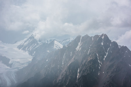 Atmospheric landscape with fuzzy silhouettes of sharp rocks and snowy mountain top in low clouds during rain. Dramatic view to large snow mountains and glacier blurred in rain haze in gray low clouds.の写真素材