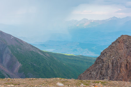Scenic view from abyss edge to green hills and sunlit mountain range in rainy clouds during rain. Dramatic landscape near precipice with mountain vastness in sunlight in clouds in changeable weather.の写真素材