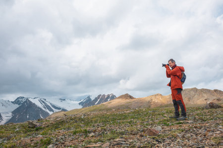 Man in red with camera shoots high snowy mountain range with sharp tops under gray cloudy sky. Tourist photographs dramatic landscape with large snow mountains with pointed peaks at changeable weatherの写真素材