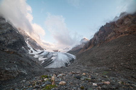 Scenic mountain landscape with large glacier in low clouds in sunrise colors. Colorful view to glacier tongue in morning sunlight in low clouds. Atmospheric high mountain scenery at early morning.の写真素材