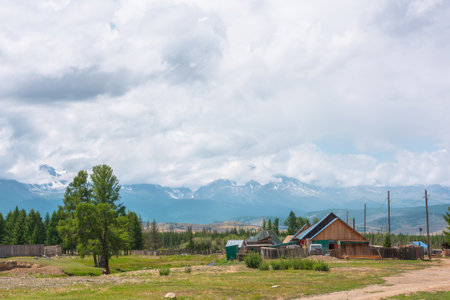 Dramatic alpine landscape of beautiful mountain village with wooden rustic houses with sloping roofs in sunlight and large mountain range in low clouds. Russia, Altai Republic, Kurai, 13 July, 2021.のeditorial素材