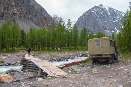 Tourist on bridge across mountain river and truck in coniferous forest with view to snow mountains. Larch trees in valley of Aktru river. Russia, Altai Republic, Aktru mountain camp, 01 July, 2021.のeditorial素材
