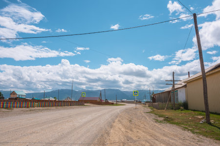 Dramatic landscape inside beautiful mountain village against large mountain range at changeable weather. Multicolor fence along sunlit road with crosswalk. Russia, Altai Republic, Kurai, 13 July, 2021のeditorial素材