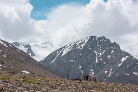 Small cabin at high altitude near precipice edge with view to high rocky mountain with peaked top. Small house against snowy pointy peak under cloudy sky. Russia, Altai Republic, Aktru, 06 July, 2021.のeditorial素材