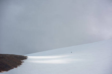 Awesome scenery with small man on large snowy mountain in sunlight under gray cloudy sky....