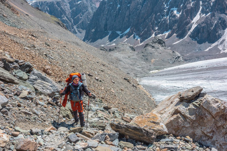 Backpacker with trekking poles walks in high mountains in sunny day. Scenic alpine landscape with tourist near glacier in sunlight. Man with sunglasses and camera among mountains with snow in sunshineの写真素材