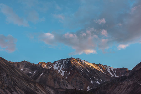 Scenic landscape with high snow mountain with sharp rocky pinnacle in golden sunlight under clouds of sunset color at changeable weather. Colorful view to large mountain top under cloudy sunset sky.の写真素材