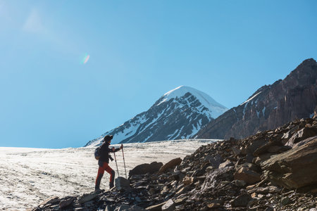 Scenic alpine landscape with silhouette of hiker with trekking poles against large glacier tongue and snow mountain peak in sunlight. Man with backpack in high mountains under blue sky in sunny day.の写真素材
