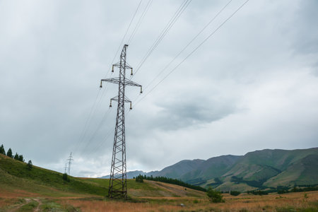 Dramatic mountain landscape with tall tower of power lines under cloudy sky. Atmospheric alpine scenery with electric tower with wires against high mountains in overcast. Electricity in mountains.の写真素材