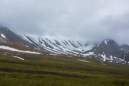 Dramatic alpine landscape with snowy mountains in gray low clouds. Bleak atmospheric scenery of tundra under lead gray sky. Gloomy minimalist view to mountain range among low rainy clouds in overcast.の写真素材