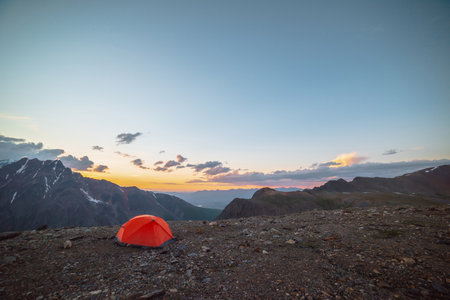Scenic alpine landscape with tent at very high altitude with view to large mountains in orange dawn sky. Vivid orange tent with awesome view to high mountain range under cloudy sky in sunset colors.の写真素材