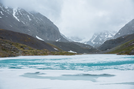 Atmospheric mountain landscape with frozen alpine lake and high snowy mountains. Awesome overcast scenery with icy mountain lake on background of snow mountains in low clouds. Scenic view to ice lake.の写真素材