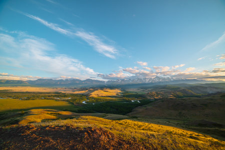 Scenic sunny view from sunlit grassy hill to forest valley with serpentine river against high snowy mountain range in sunlight. Beautiful snake mountain river in forest and snow mountains in sunshine.の写真素材