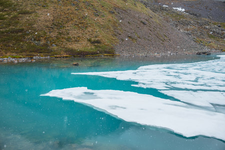 Atmospheric mountain landscape with frozen alpine lake during snowfall. Clear ice floats on water surface of lake. Awesome mountain scenery with icy mountain lake. Flakes of snow on water background.の写真素材