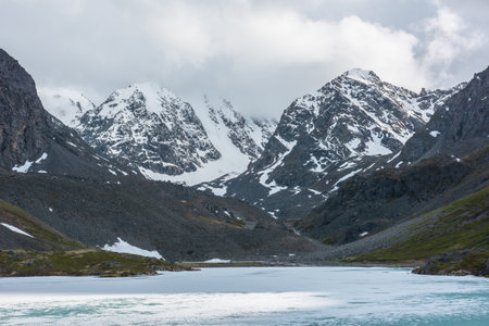Dramatic mountain landscape with frozen alpine lake and high snow mountains and sunlit big rocks among low clouds. Atmospheric scenery with ice lake on background of large snowy mountains in overcast.の写真素材