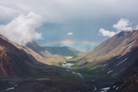 Bleak alpine landscape with vivid rainbow above mountain lake in valley. Gloomy scenery with bright rainbow above lake in mountain valley. Top view to colorful rainbow and low clouds in mountains.の写真素材