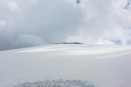 Dramatic alpine landscape with huge snowy mountain dome with rocky top in sunlight under gray cloudy sky. Awesome mountain scenery with high snow mountain in dome shape in center at changeable weatherの写真素材
