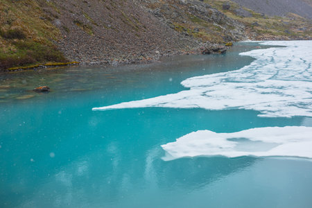 Atmospheric mountain landscape with frozen alpine lake during snowfall. Clear ice floats on water surface of lake. Awesome mountain scenery with icy mountain lake. Flakes of snow on water background.の写真素材