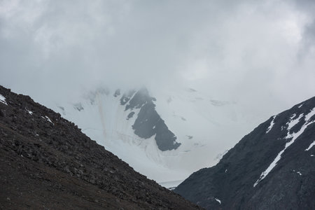 Atmospheric landscape with silhouettes of sharp rocks and snowy mountain wall in low clouds during rain. Dramatic bleak view to large snow mountains and glacier blurred in rain haze in gray low cloudsの写真素材