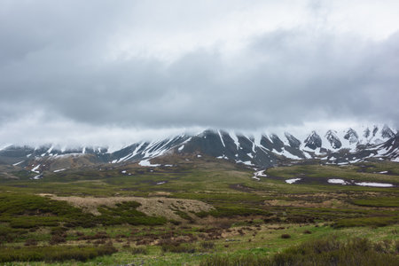 Dramatic alpine landscape with snowy mountains in gray low clouds. Bleak atmospheric scenery of tundra under lead gray sky. Gloomy minimalist view to mountain range among low rainy clouds in overcast.の写真素材