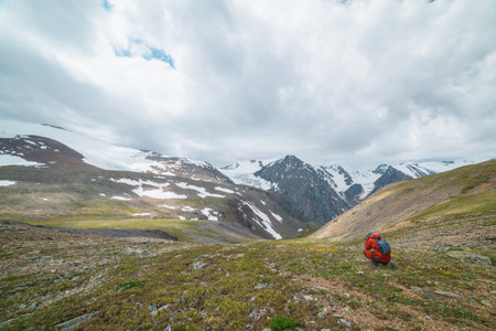 Man in red with camera shoots view with high snowy mountain range with sharp tops and glaciers under gray cloudy sky. Tourist photographs landscape with large snow mountains at changeable weather.の写真素材